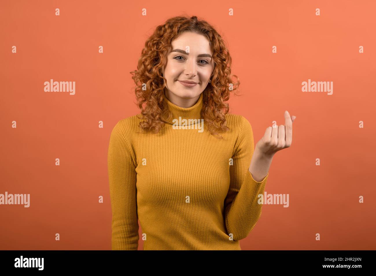 Attraktive junge kaukasische rothaarige Mädchen in orange casual Jumper zeigt Geld Geste mit Hand isoliert auf Studio orange Hintergrund. Stockfoto