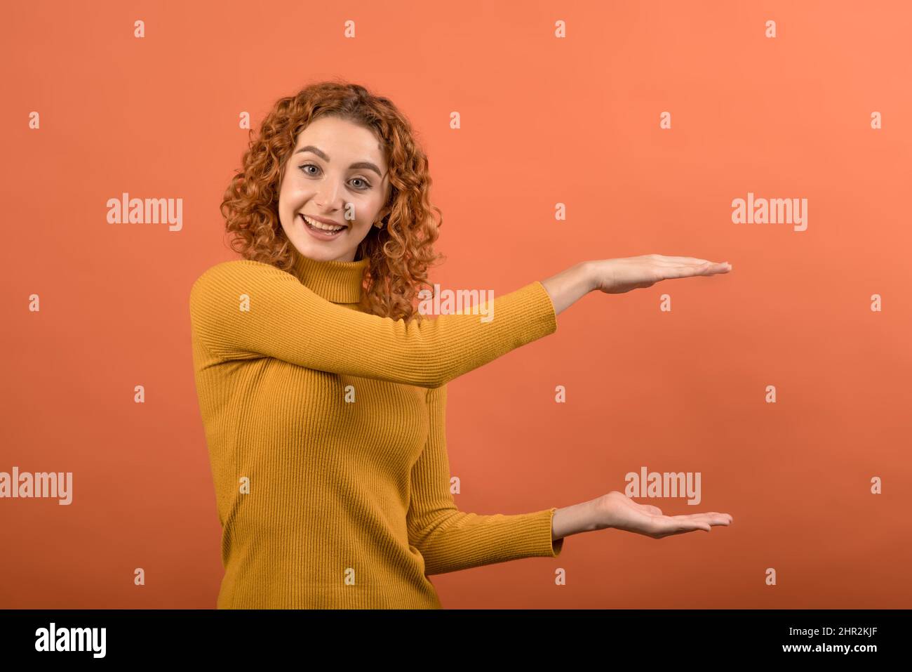 Ein junges und attraktives kaukasisches Rotschopf-Mädchen in einem orangefarbenen Pullover, das mit ihren Händen ein großes Werbeobjekt zeigt, das auf einer Orange isoliert ist Stockfoto