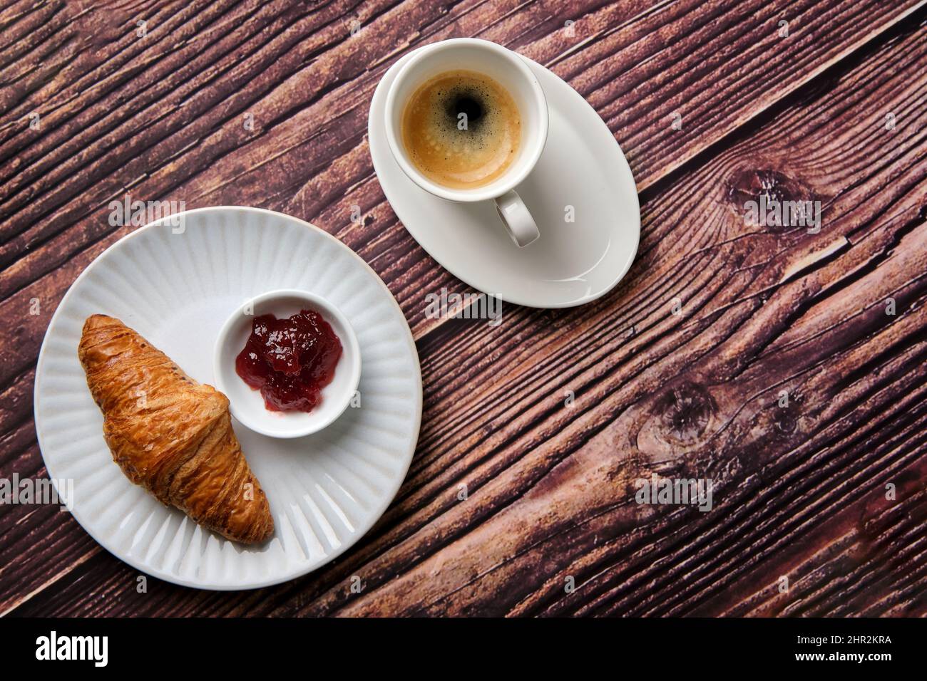 Ein Croissant, Erdbeermarmelade & Espresso-Kaffee auf einem weißen, strukturierten runden Teller auf einem Holzhintergrund von oben, ein Frühstücksfavorit weltweit. Stockfoto