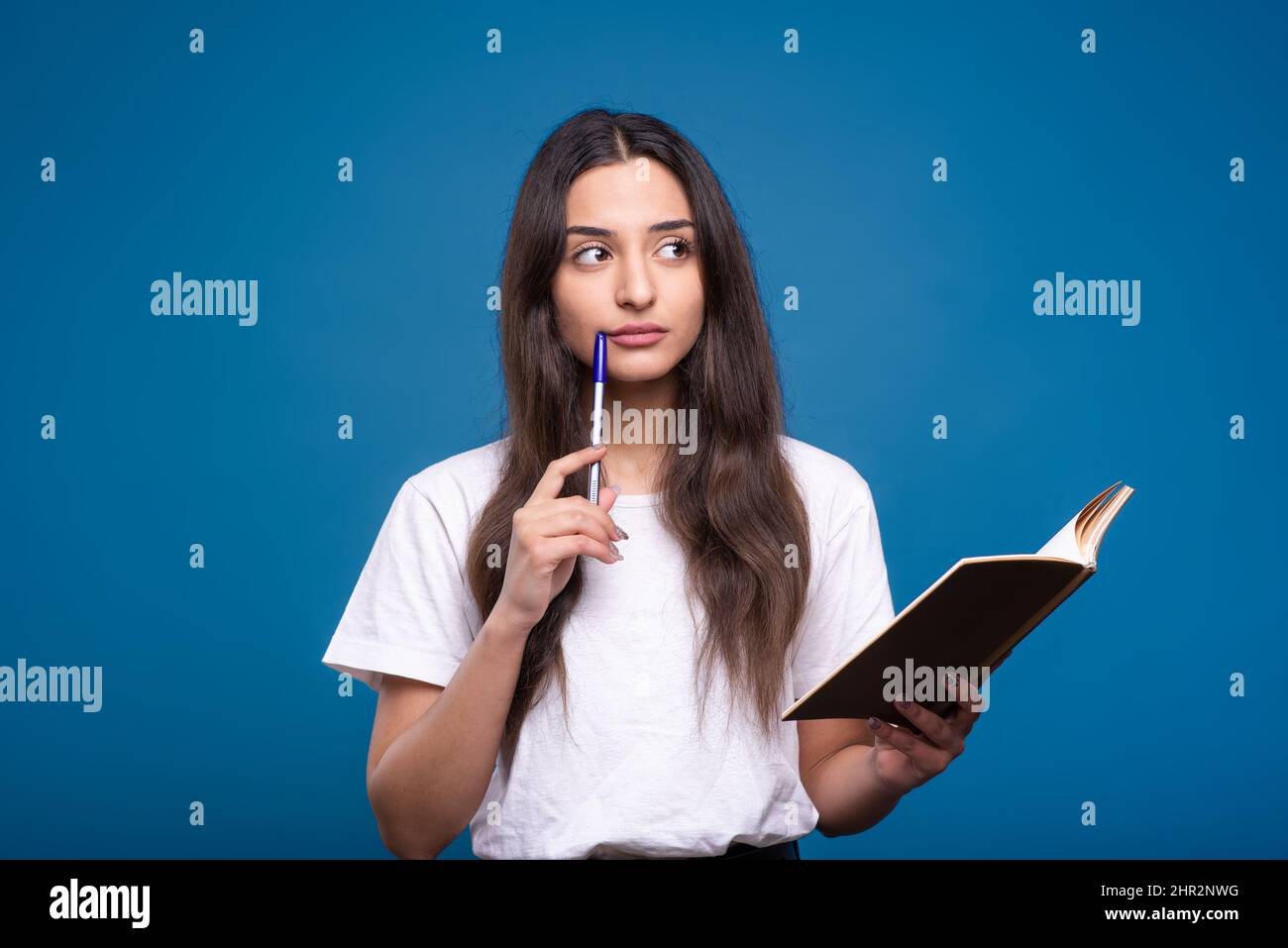 Attraktive kaukasische oder arabische Brünette Mädchen in einem weißen T-Shirt mit einem Notizbuch, Schreiben und Denken isoliert auf einem blauen Studio-Hintergrund. Stockfoto