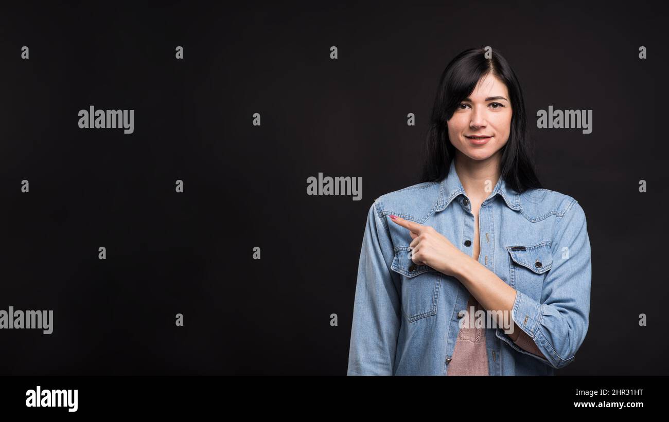 Attraktive kaukasische Brünette Mädchen in einem Hemd zeigt einen Finger neben einem Kopierplatz für Werbung isoliert auf einem schwarzen Studio Hintergrund. Stockfoto