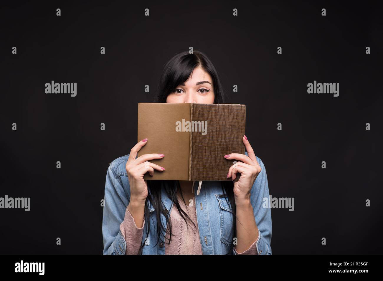 Attraktive kaukasische Brünette Mädchen in einem Hemd bedeckt ihr Gesicht mit einem Buch und Lesung isoliert auf einem schwarzen Studio-Hintergrund. Stockfoto