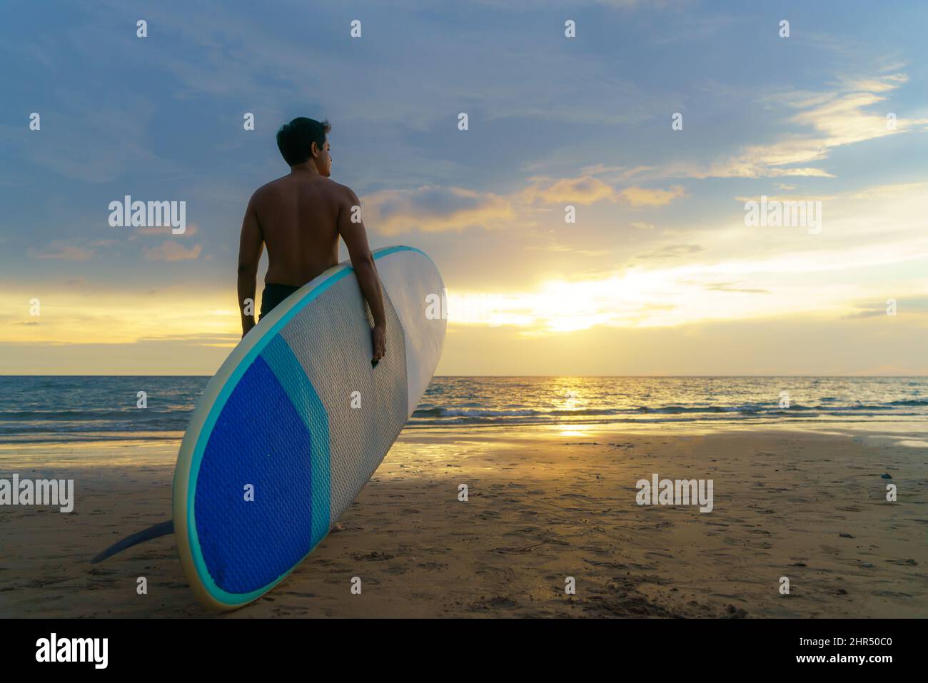 Asiatischer Mann hält ein Brett und paddelt und läuft am Strand. Im Hintergrund das Meer und der Sonnenuntergang. Rückansicht. Surfen im Sommer. Stockfoto