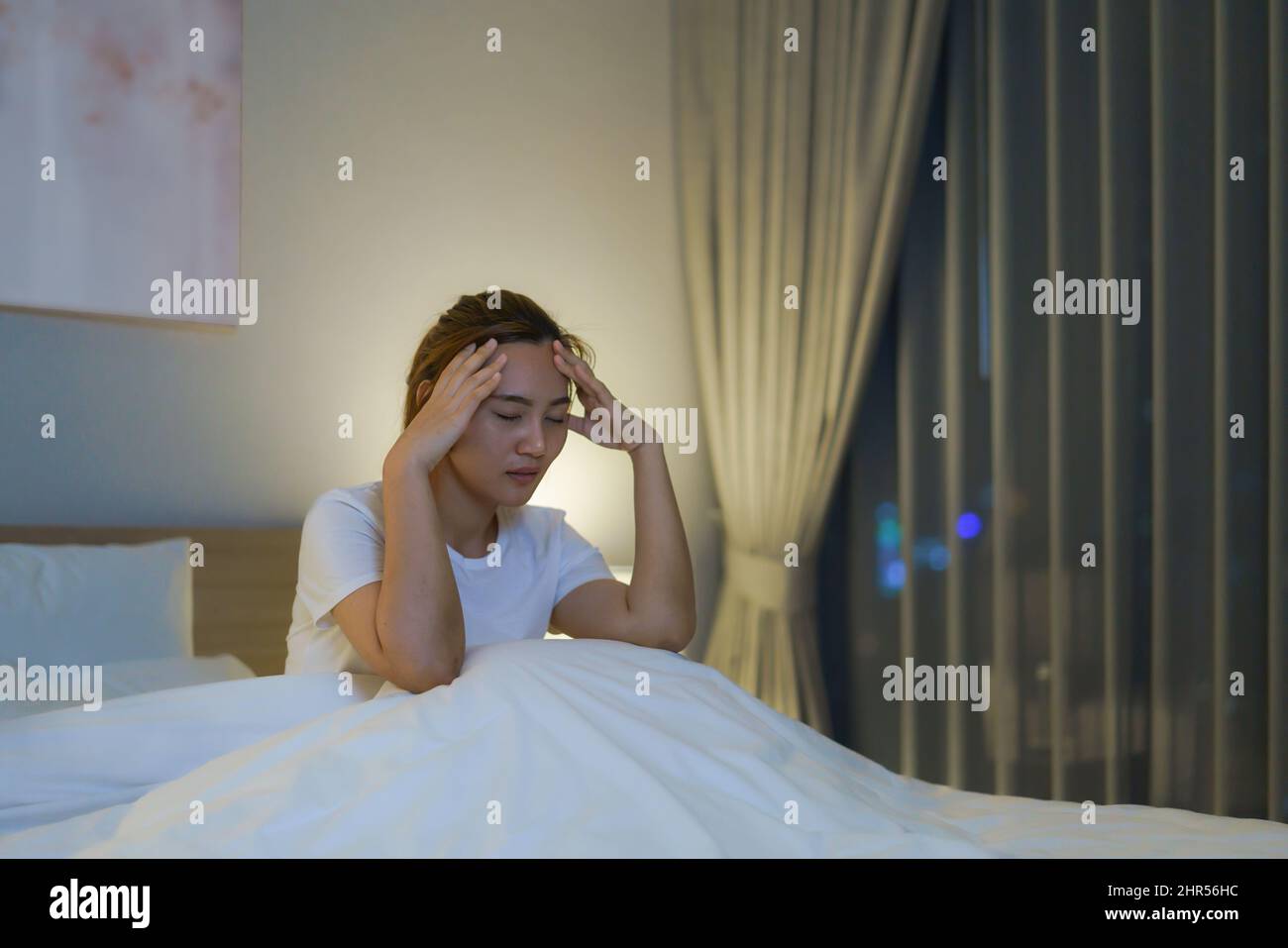 Asiatische Frau, die auf einem weißen Bett im Schlafzimmer liegt und besorgt aussieht oder über etwas über ihr Leben oder ihre Arbeit in der Nacht zu Hause nachdenkt. Stockfoto
