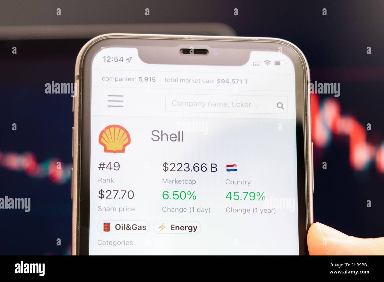 Shell-Aktienkurs auf dem Bildschirm des Mobiltelefons in mans Hand mit wechselnden Börsengraphen im Hintergrund, Februar 2022, San Francisco, USA. Stockfoto