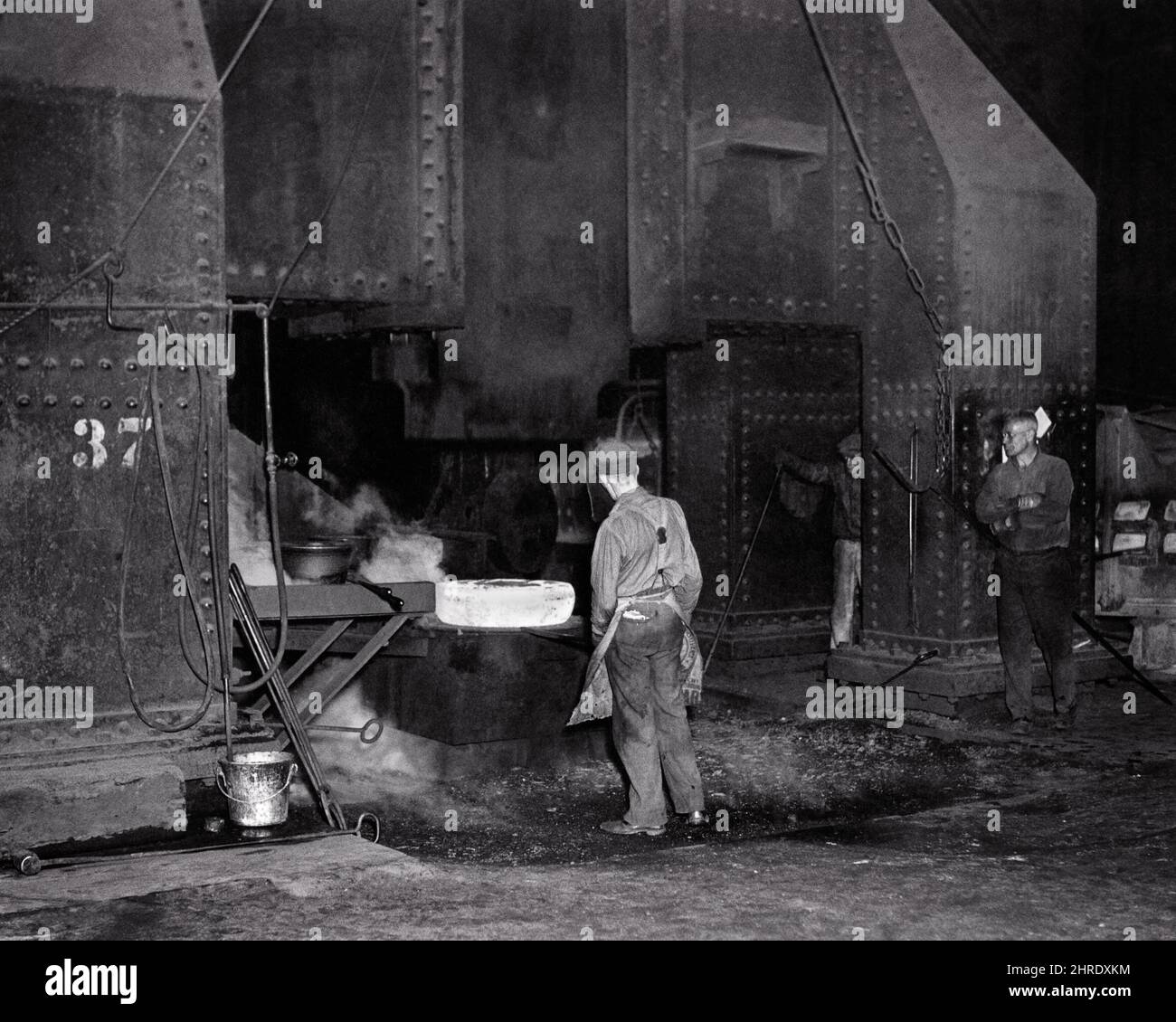 1930S MÄNNER PRODUKTIONSARBEITER BEOBACHTEN RIESIGE PRESSE STAMPFEN ROT-HEISSEN STAHL EISENBAHNWAGEN RÄDER STAHLWERK CHESTER PA USA - I202 HAR001 HARS VEREINIGTE STAATEN VON AMERIKA SCHWERE MÄNNER RISIKO BERUF RÄDER B&W NORDAMERIKA NORTH AMERICAN SKILL BERUF FÄHIGKEITEN UND KARRIERE WISSEN PA ARBEIT IN BESCHÄFTIGUNG FERTIGUNG BERUFE STANDARD KONZEPTUELLE WERKE FACILITY INFRASTRUKTUR CASTING MITARBEITER RIESIG MITTLEREN ERWACHSENEN MITTLEREN ERWACHSENEN MANN ROT-HEISS SCHWARZ UND WEISS KAUKASISCHEN ETHNIZITÄT HAR001 ARBEIT ALTMODISCH Stockfoto