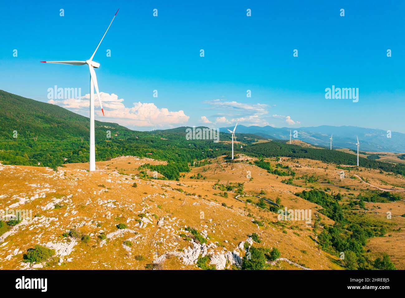 Windmühlen mit großen Propellern produzieren grüne Energie im Hochland gegen forstwirtschaftliche Berge unter blauem Himmel bei einem sonnigen Sommertag aus der Luft Stockfoto