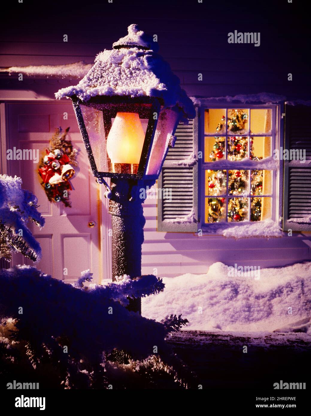 1970S WEIHNACHTEN OUTDOOR NACHT SZENE SCHNEE BEDECKT WILLKOMMEN LAMPPOST LICHT UND VERSCHALT FENSTER MIT DEKORIERTEN BAUM SICHTBAR INNEN - KX7330 PHT001 HARS AUSSEN DEZEMBER KONZEPTIONELL DEZEMBER 25 STILLLEBEN STILVOLL SICHTBAR FREUDIG LAMPPOST GESCHLOSSEN ZWEISAMKEIT ALTMODISCH Stockfoto