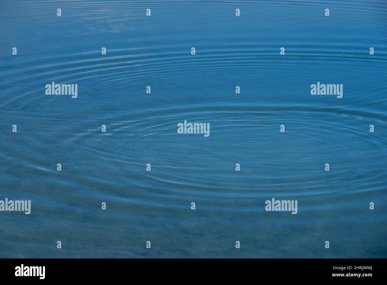 Hintergrund von der Oberfläche eines Wasserkörpers, auf dem sich kleine Wellen im Kreis ausbreiten, im blauen Wasser Stockfoto