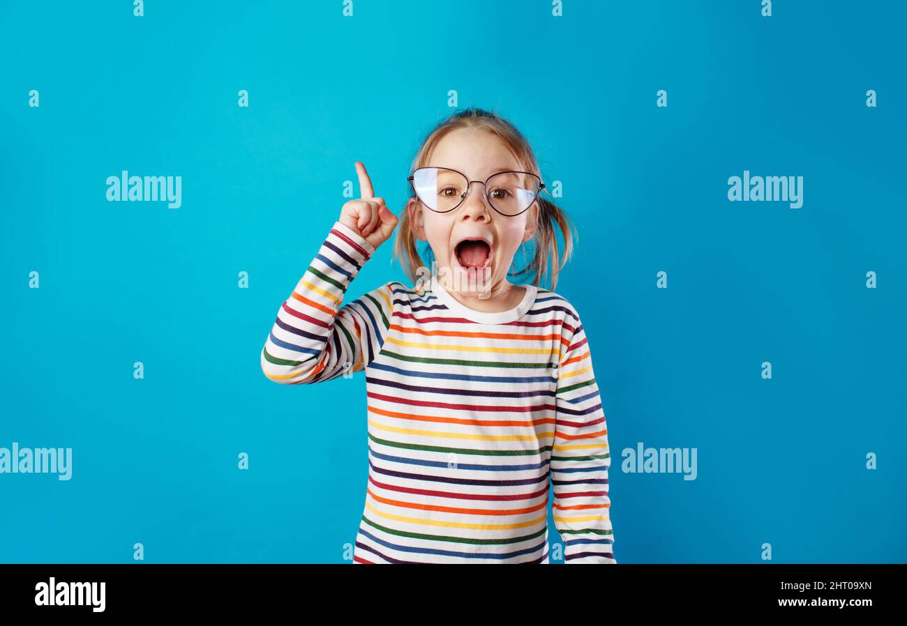 Ein kleines Mädchen mit großen Brillen und einem gestreiften T-Shirt steht auf einem blauen, isolierten Hintergrund mit weit offenem Mund. Stockfoto