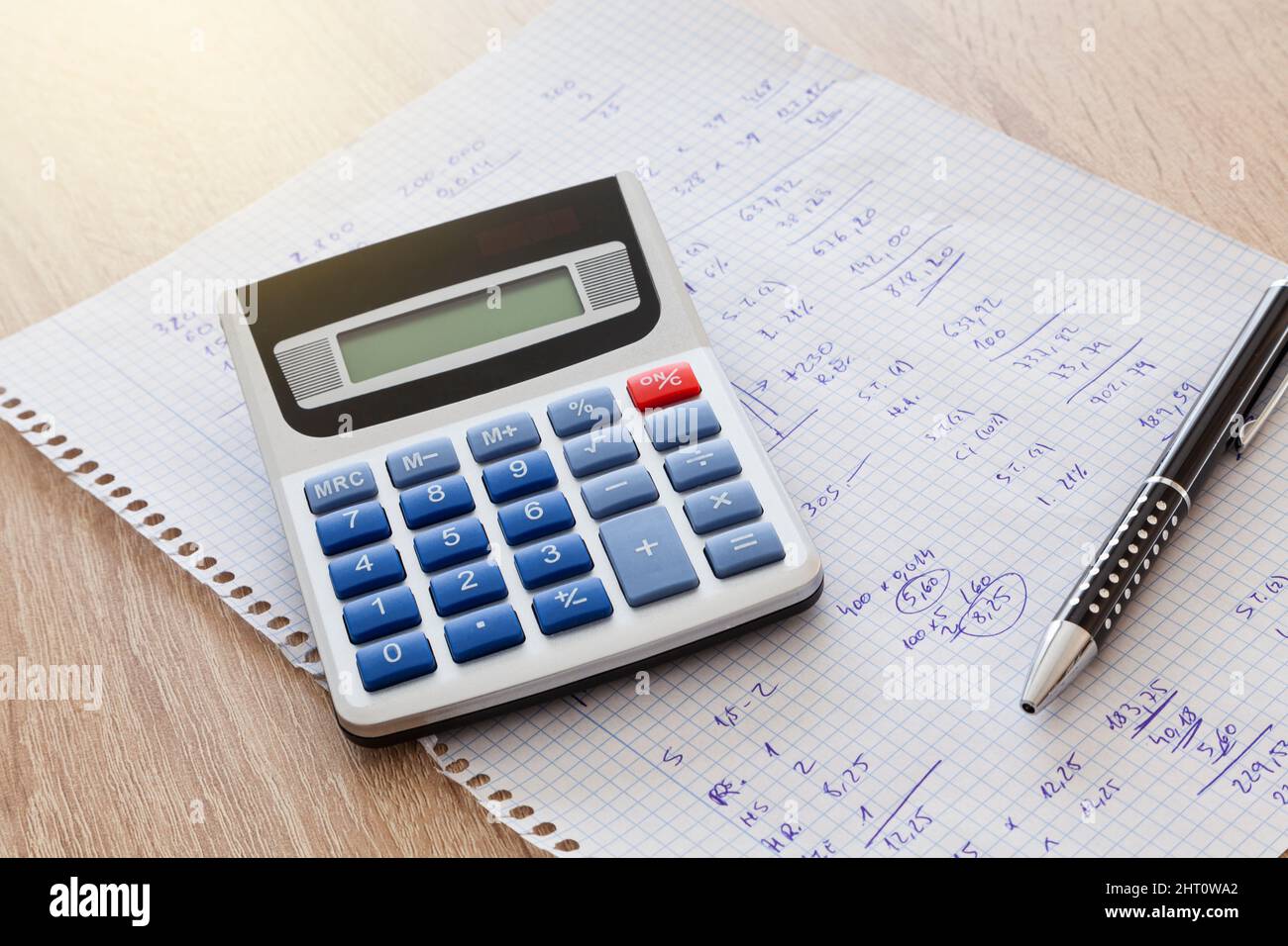 Auf einem Holztisch befindet sich ein Blatt Notizbuchquadrate mit handschriftlichen Zahlen und Berechnungen und auf dem Blatt ist ein Stift und ein Rechner. Stockfoto