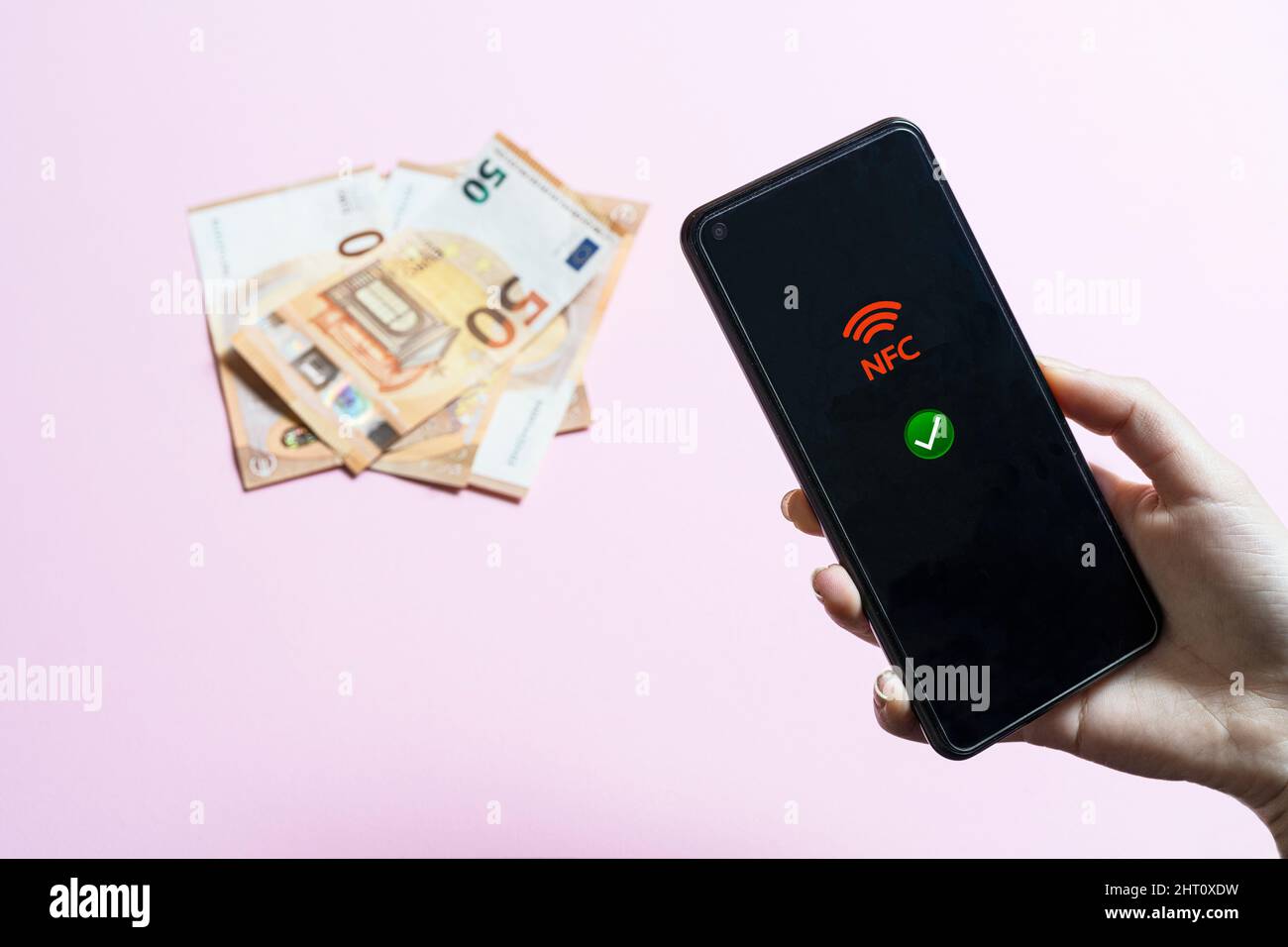 Das Mädchen hält ein Handy in der Hand, das NFC-Symbol auf dem Bildschirm und einige Euro-Banknoten im Hintergrund Stockfoto