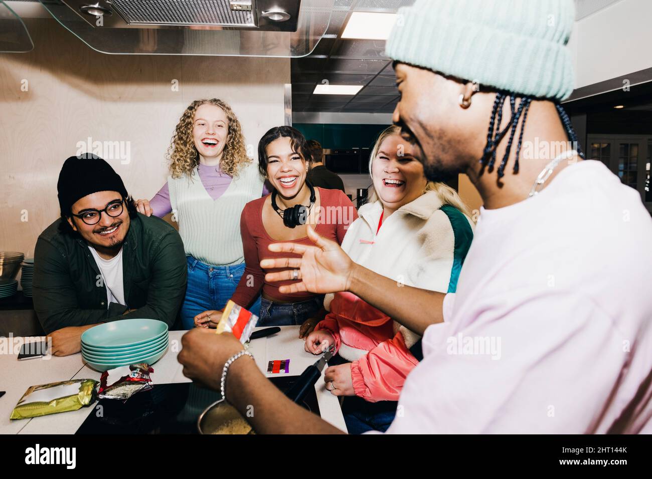 Glücklicher multirassischer junger Mann und Frauen, die einen männlichen Freund bei der Zubereitung von Essen im Studentenwohnheim anguckt Stockfoto