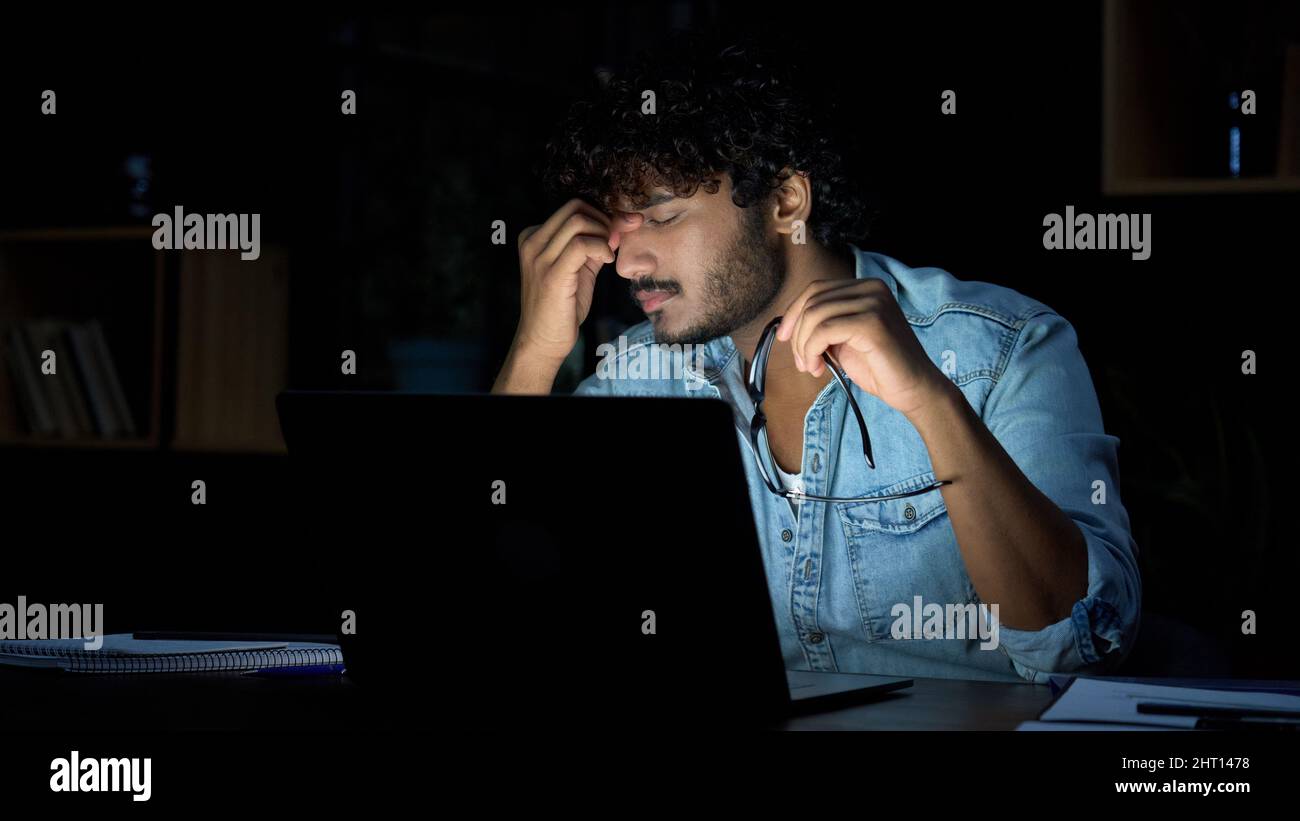 Überarbeiteter junger indischer Geschäftsmann, der spät am Arbeitsplatz eine Brille auszieht. Stockfoto