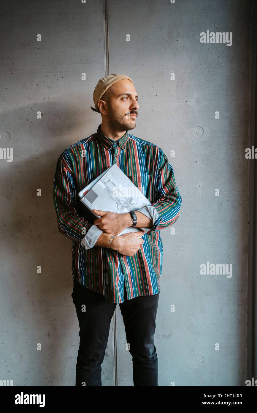 Nachdenklicher Student mit einem Buch, der vor einer grauen Wand an der Hochschule wegschaut Stockfoto