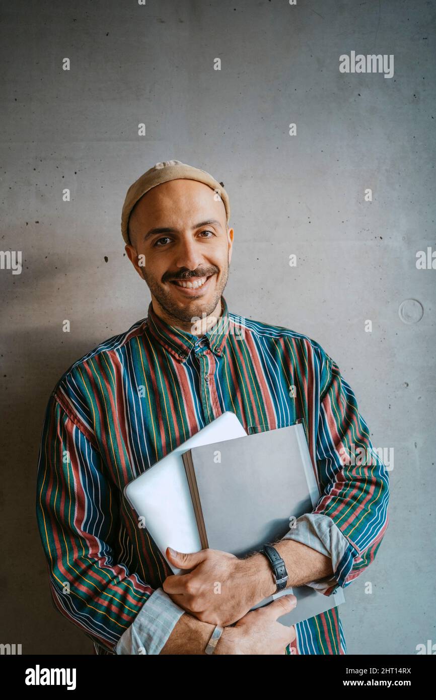 Porträt eines lächelnden männlichen erwachsenen Studenten, der ein Buch vor einer grauen Wand an der Hochschule hält Stockfoto