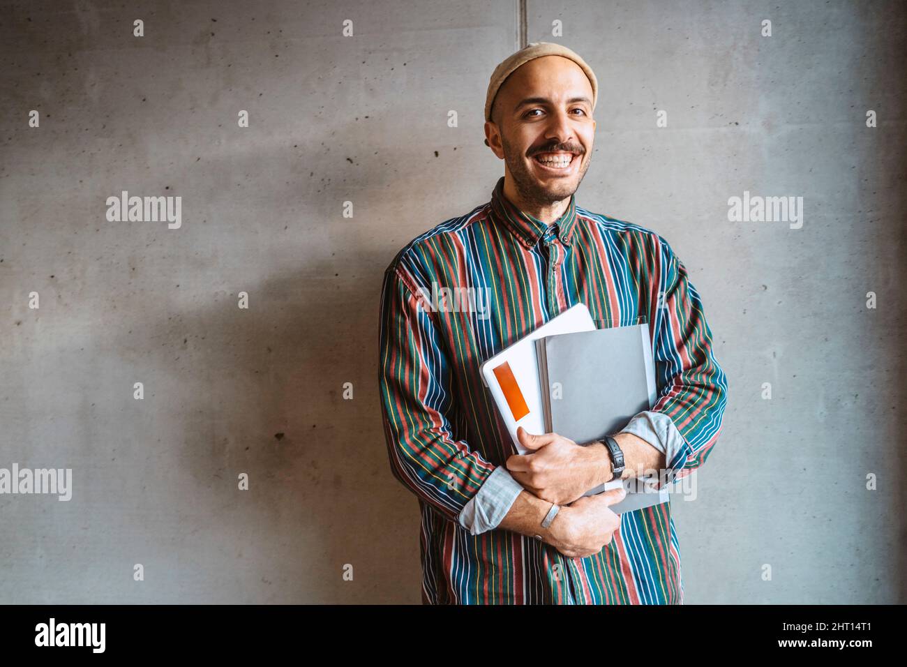 Porträt eines fröhlichen männlichen erwachsenen Studenten, der ein Buch vor einer grauen Wand an der Hochschule hält Stockfoto