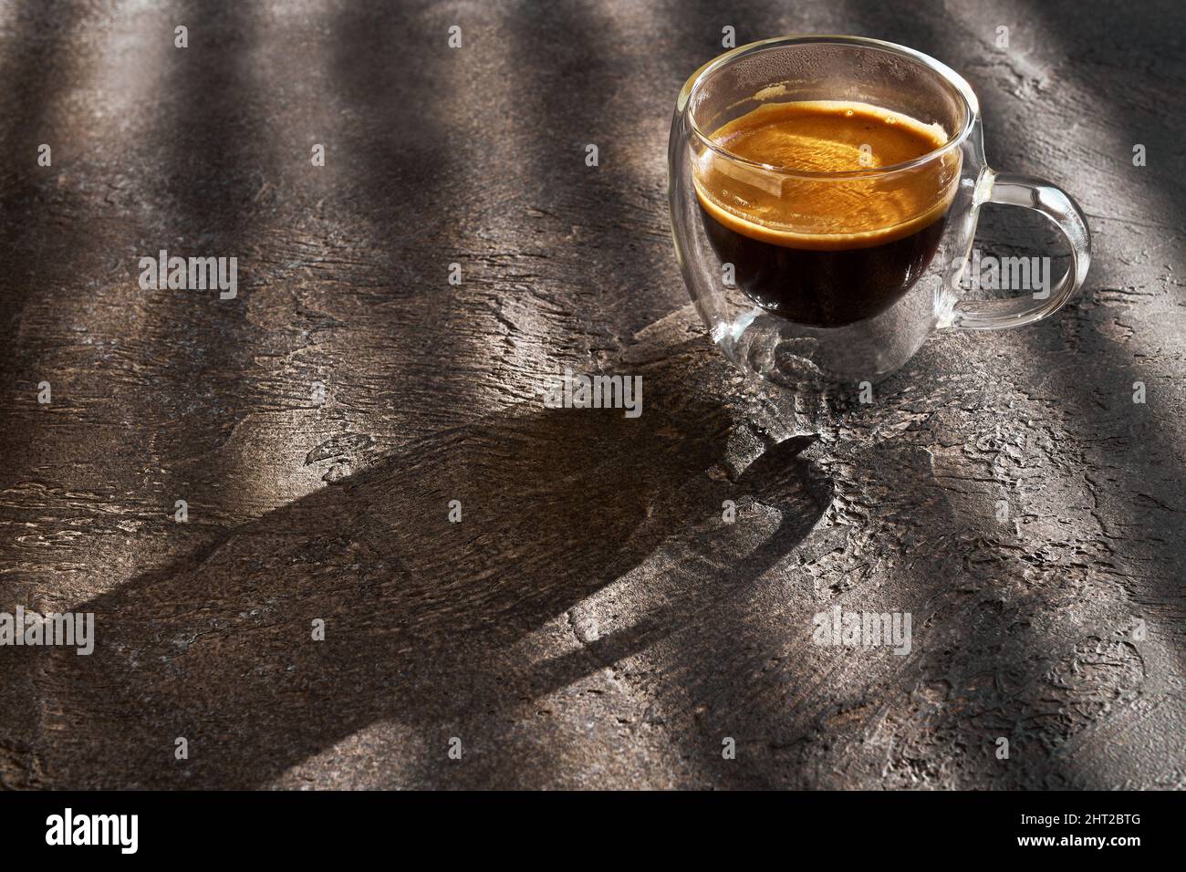Glas-Tasse mit doppeltem Espresso am Morgen bei Sonnenlicht auf einem Tisch mit dunkler Struktur, Nahaufnahme, geringe Schärfentiefe Stockfoto