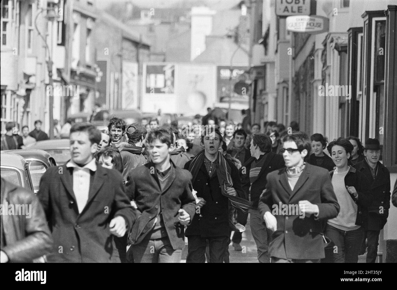 Die Mods und Rockers waren zwei widersprüchliche britische Jugendsubkulturen von Anfang bis Mitte 1960s. Die Berichterstattung über die Kämpfe von Mods und Rockern im Jahr 1964 löste eine moralische Panik über britische Jugendliche aus, und die beiden Gruppen wurden als Volksteufel bezeichnet. Mods, die durch die Straßen von Brighton laufen. 18.. April 1965. Stockfoto