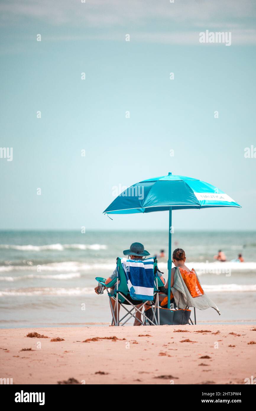 Eine vertikale Aufnahme junger Frauen, die unter einem blauen Sonnenschirm am Strand sitzen Stockfoto