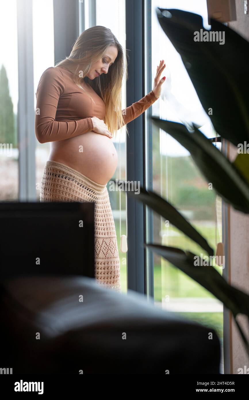 Die junge blonde, schwanger gebliebene Frau blickt auf ihren Bauch nahe dem Fenster. Stockfoto