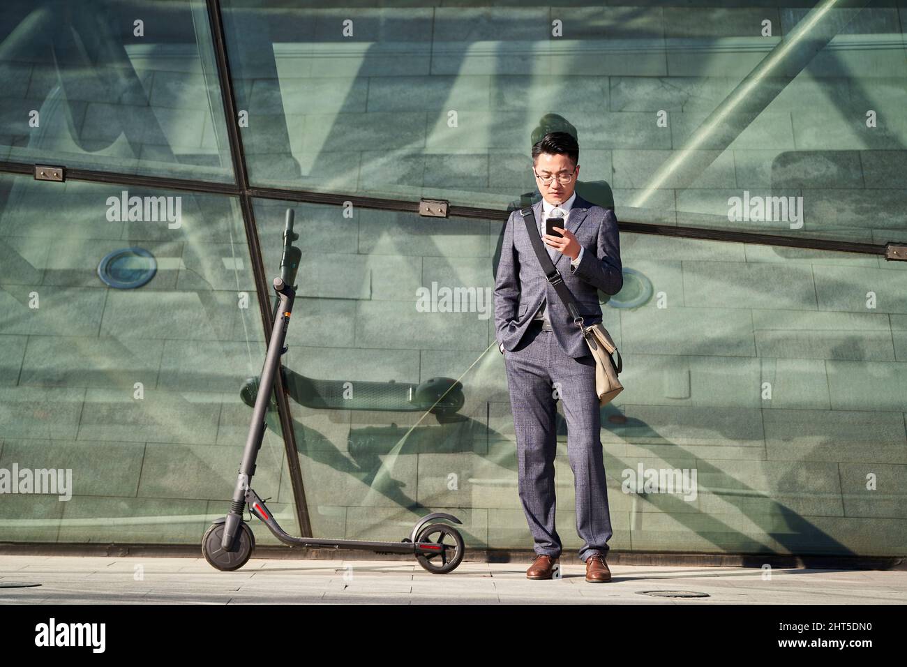 Junge asiatische Geschäftsmann mit Elektroroller Blick auf Handy im Freien Stockfoto