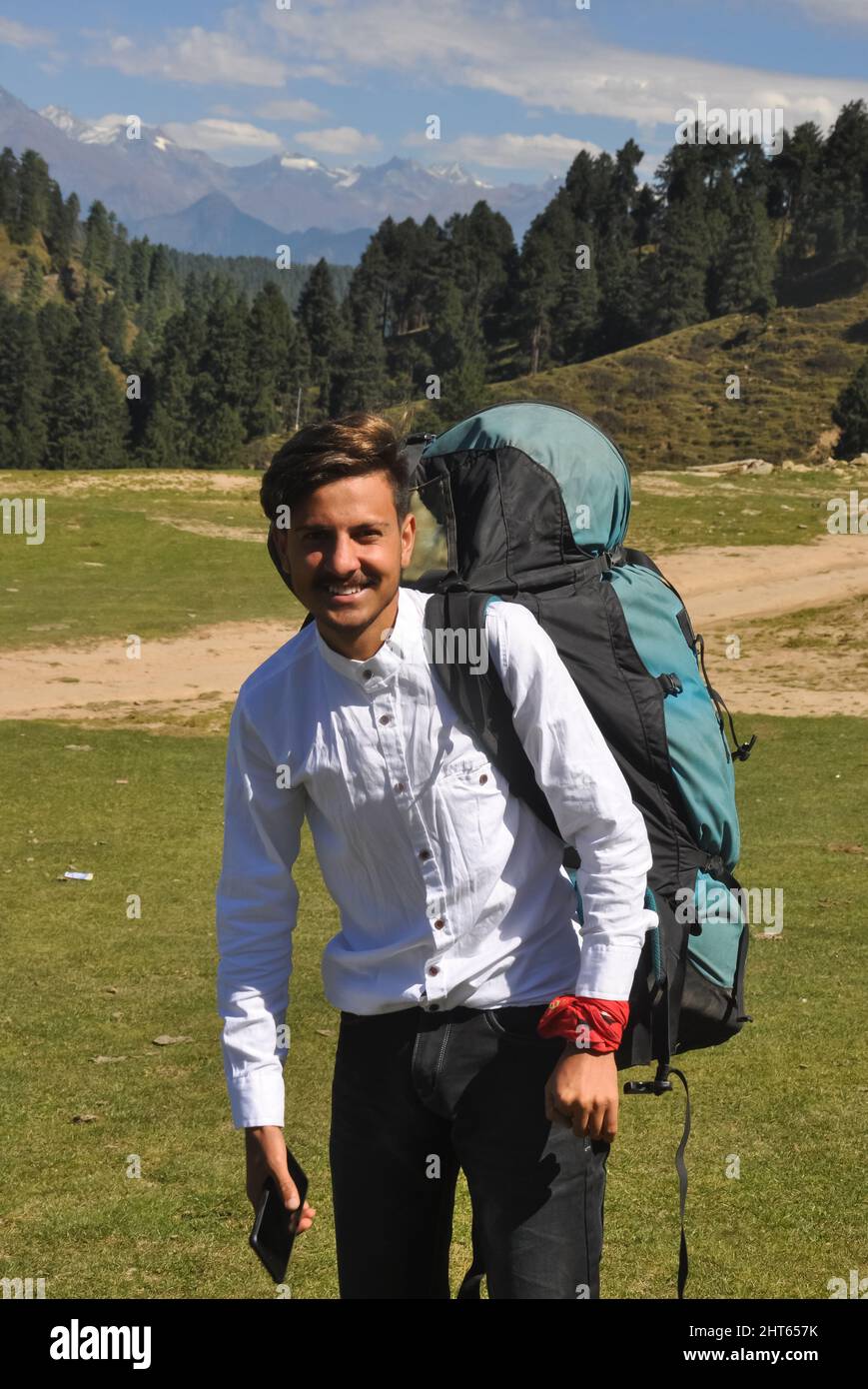 Vorderansicht eines glücklichen jungen Mannes, der die Kamera ansieht und einen Fallschirmrucksack im Berg trägt Stockfoto
