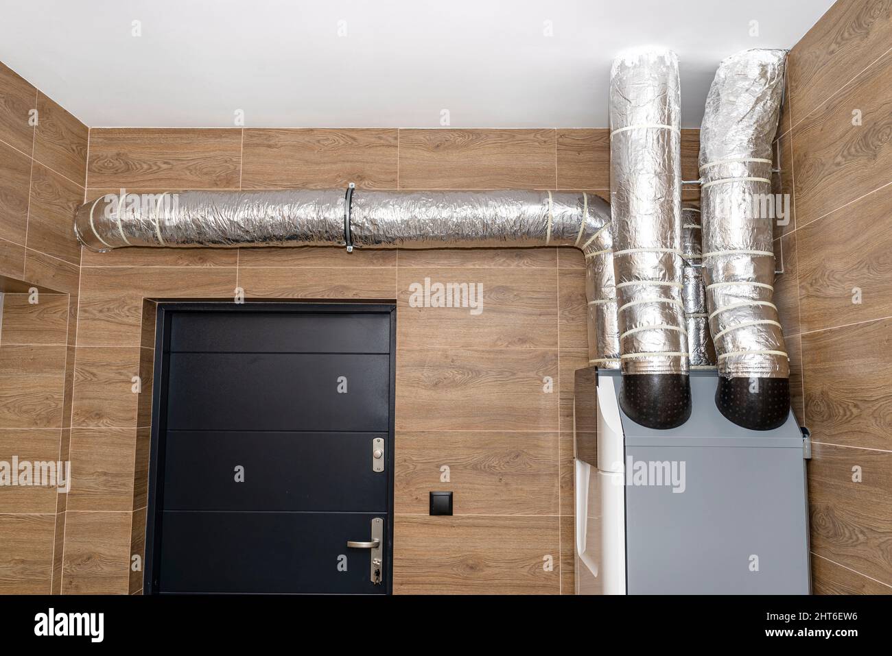 Mechanische Belüftung mit Wärmerückgewinnung, die an der Wand in einem modernen Gaskessel-Raum mit braunen Keramikfliesen hängt, die Holz imitieren. Stockfoto