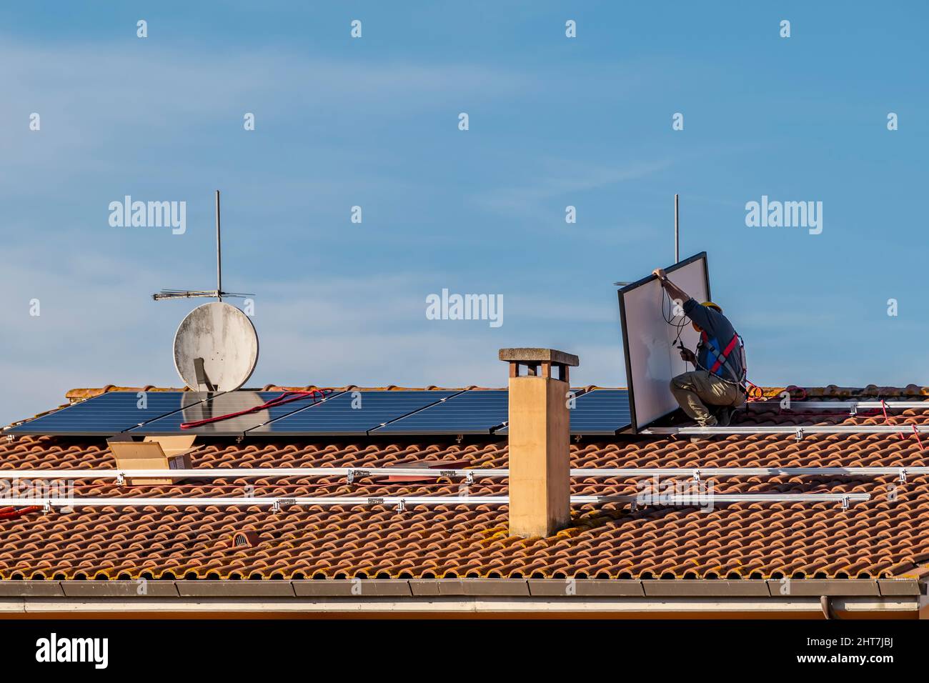 Ein Techniker hebt eine Solaranlage an, während er eine Photovoltaikanlage auf einem rot gefliesten Dach installiert Stockfoto