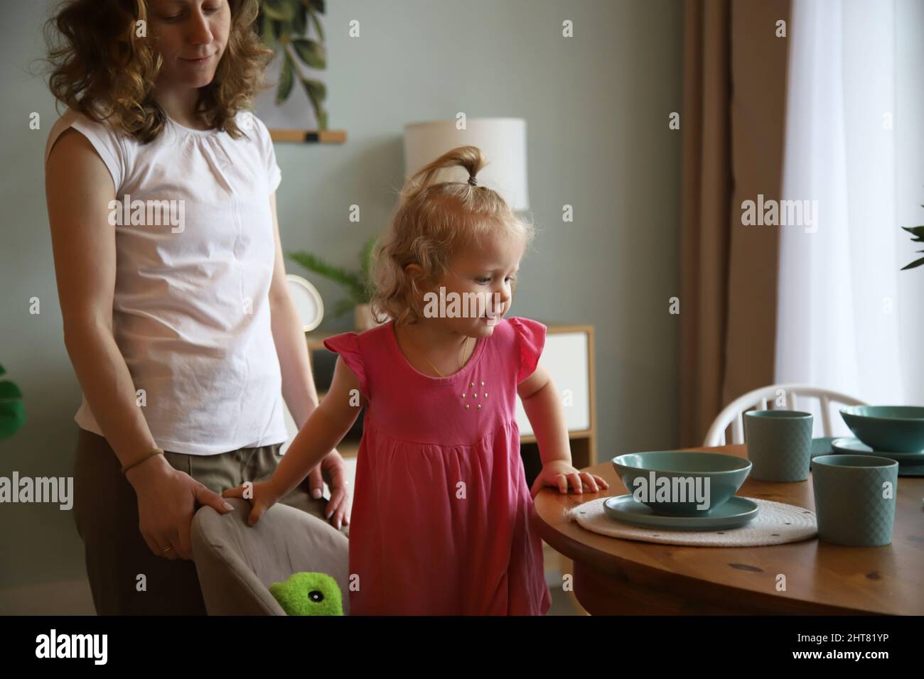 Mama hilft dem Baby, sich an den Tisch zu setzen Stockfoto