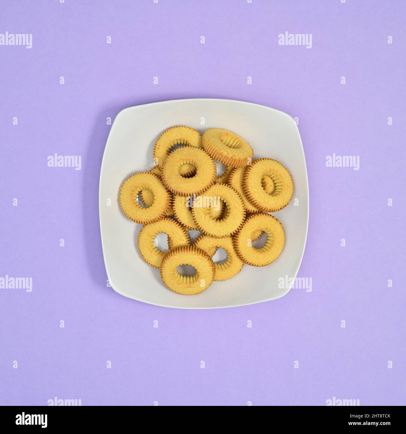 Süße Kekse in weißem Teller auf pastellviolettem Hintergrund. Flat-Lay-Food-Konzept Stockfoto