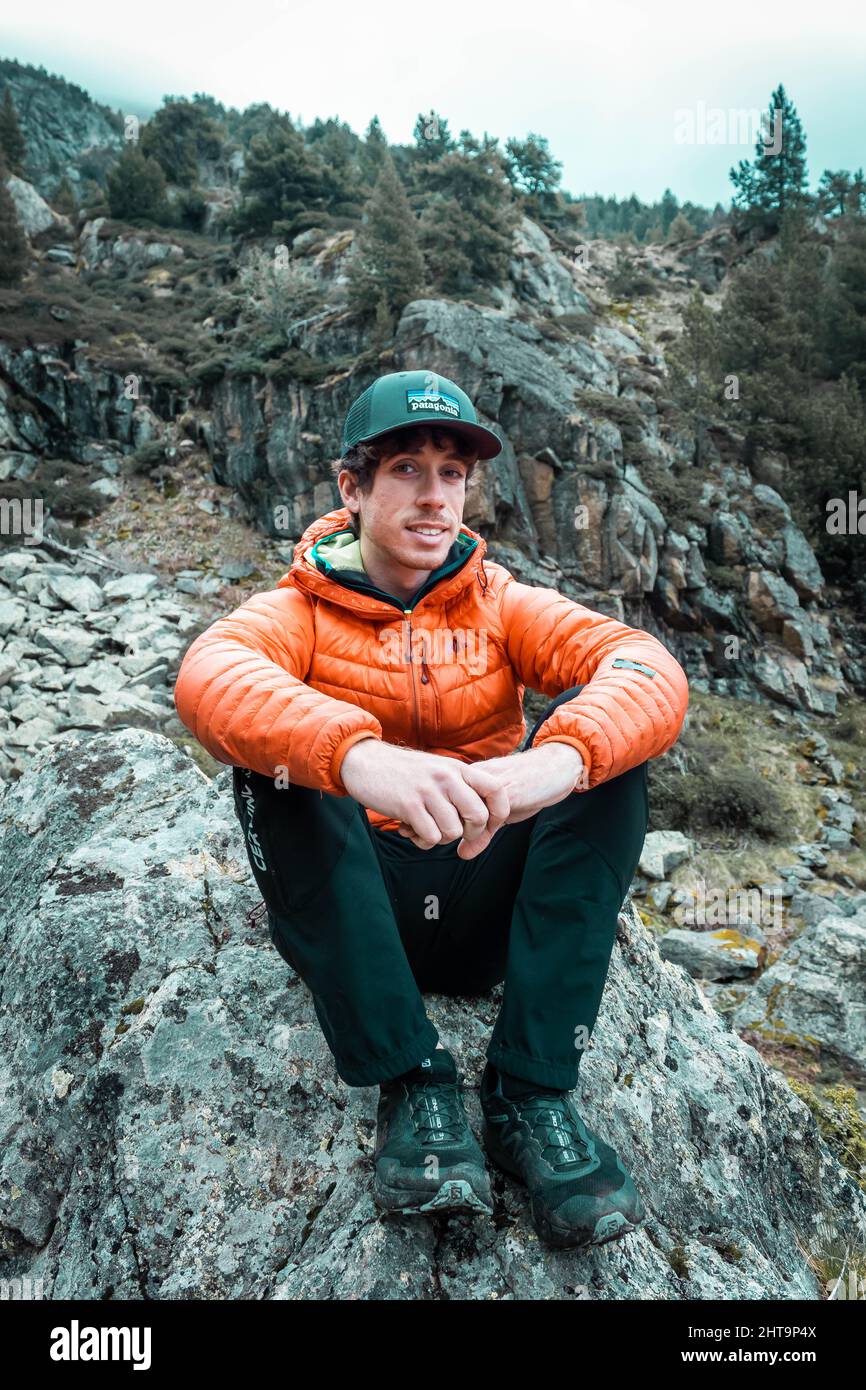 Eine vertikale Aufnahme einer Person, die sich in Andorra in einem Bergpfad ausruhte und eine orangefarbene Jacke und einen grauen Hut trug Stockfoto