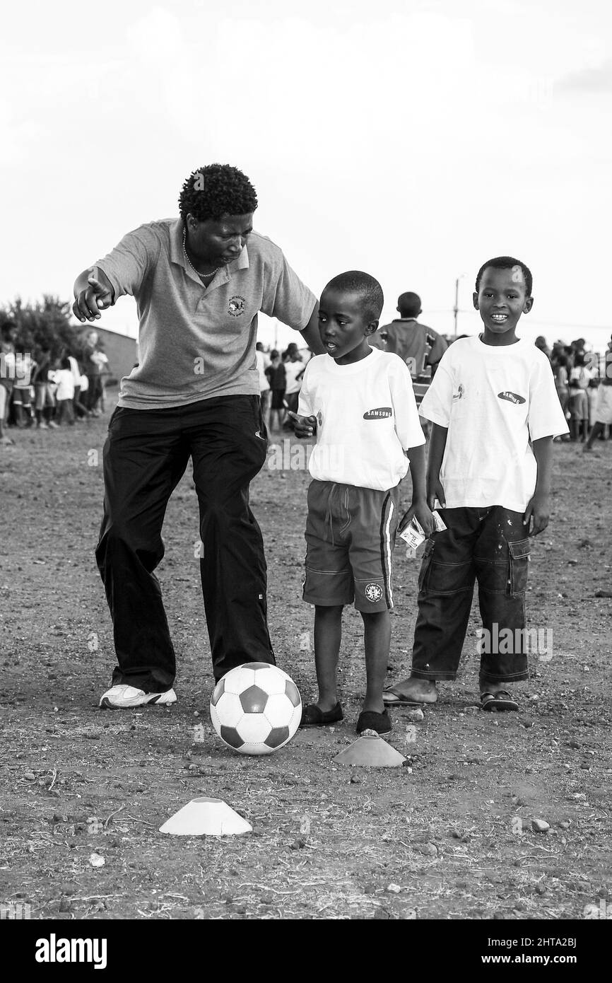 Eine Graustufenaufnahme eines freiwilligen Fußballtrainers, der mit jungen afrikanischen Kindern auf dem Schulhof arbeitet Stockfoto