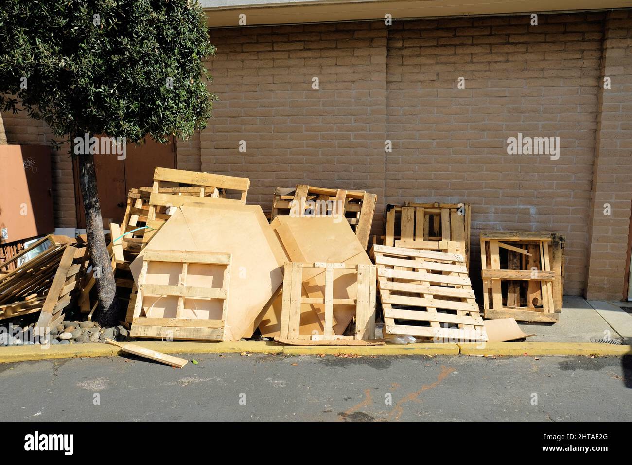 Verschiedene Holzpaletten und andere Verpackungsmaterialien aus Holz für den Transport von Geräten und Fertigwaren auf einem Bürgersteig an Wand, Baum. Stockfoto