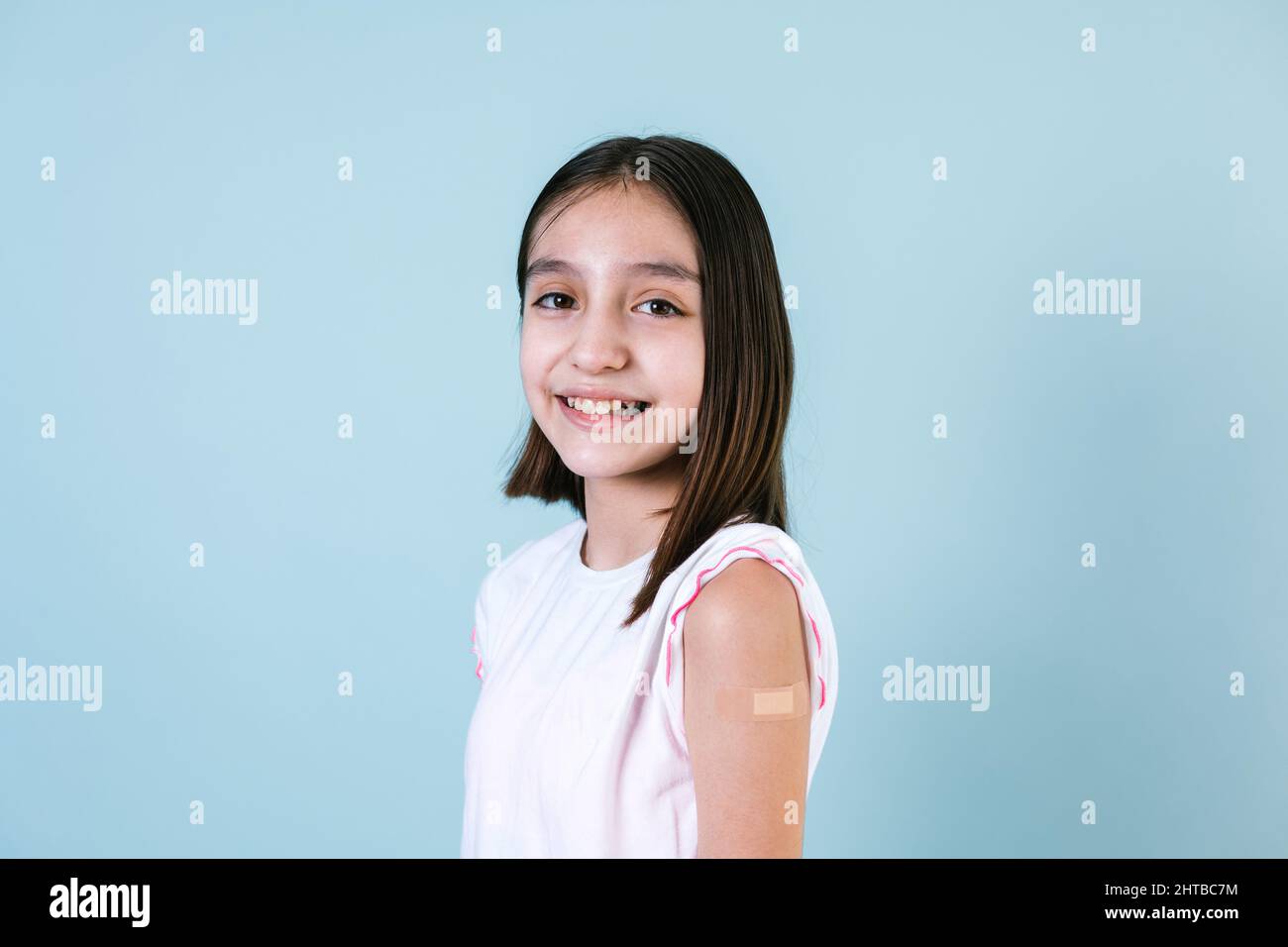 Hispanische Mädchen Kinderportrait, nachdem sie einen Impfschutz erhalten hatte und ihren Arm mit einem Verband gezeigt hatte, der in Mexiko La eine Impfung erhielt, auf blauem Hintergrund Stockfoto