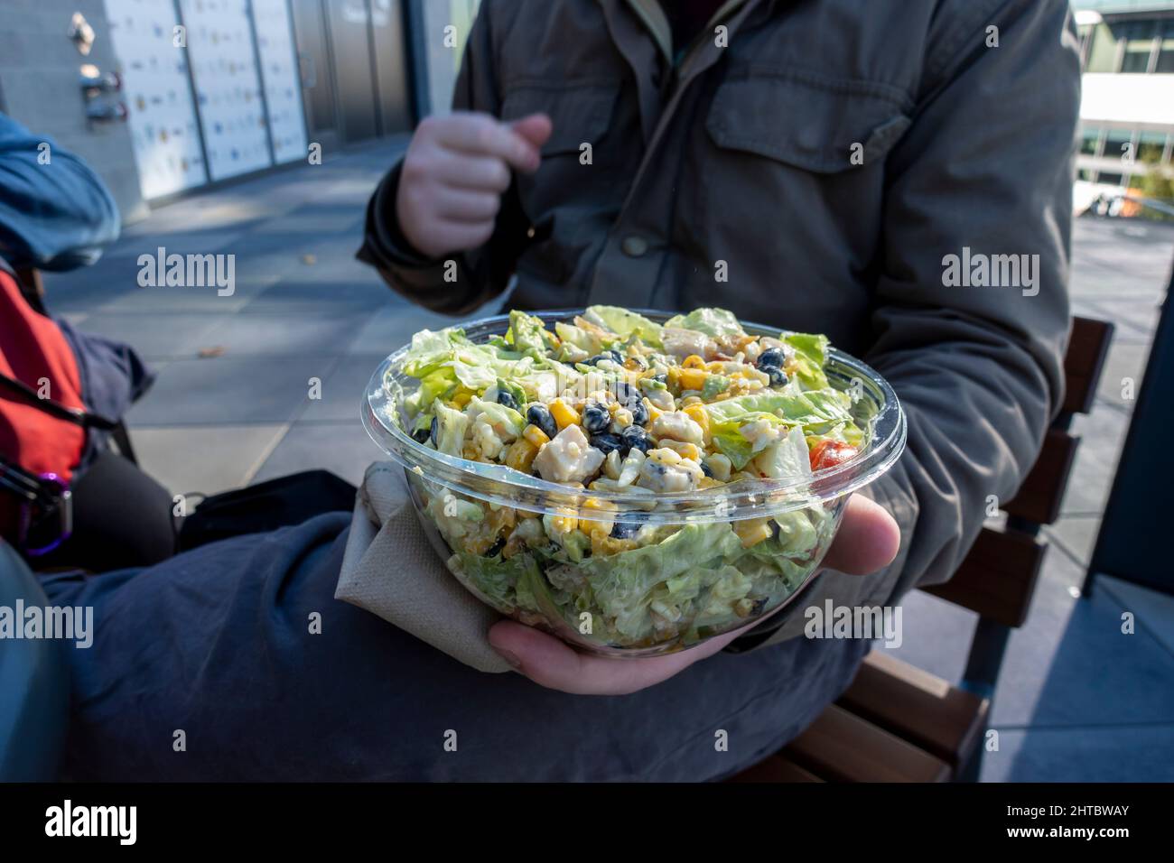 Kaukasisches Männchen in einer Winterjacke, das einen südwestlichen Salat in einem Plastikbehälter zum Mitnehmen an einem Tisch im Freien aushält. Stockfoto