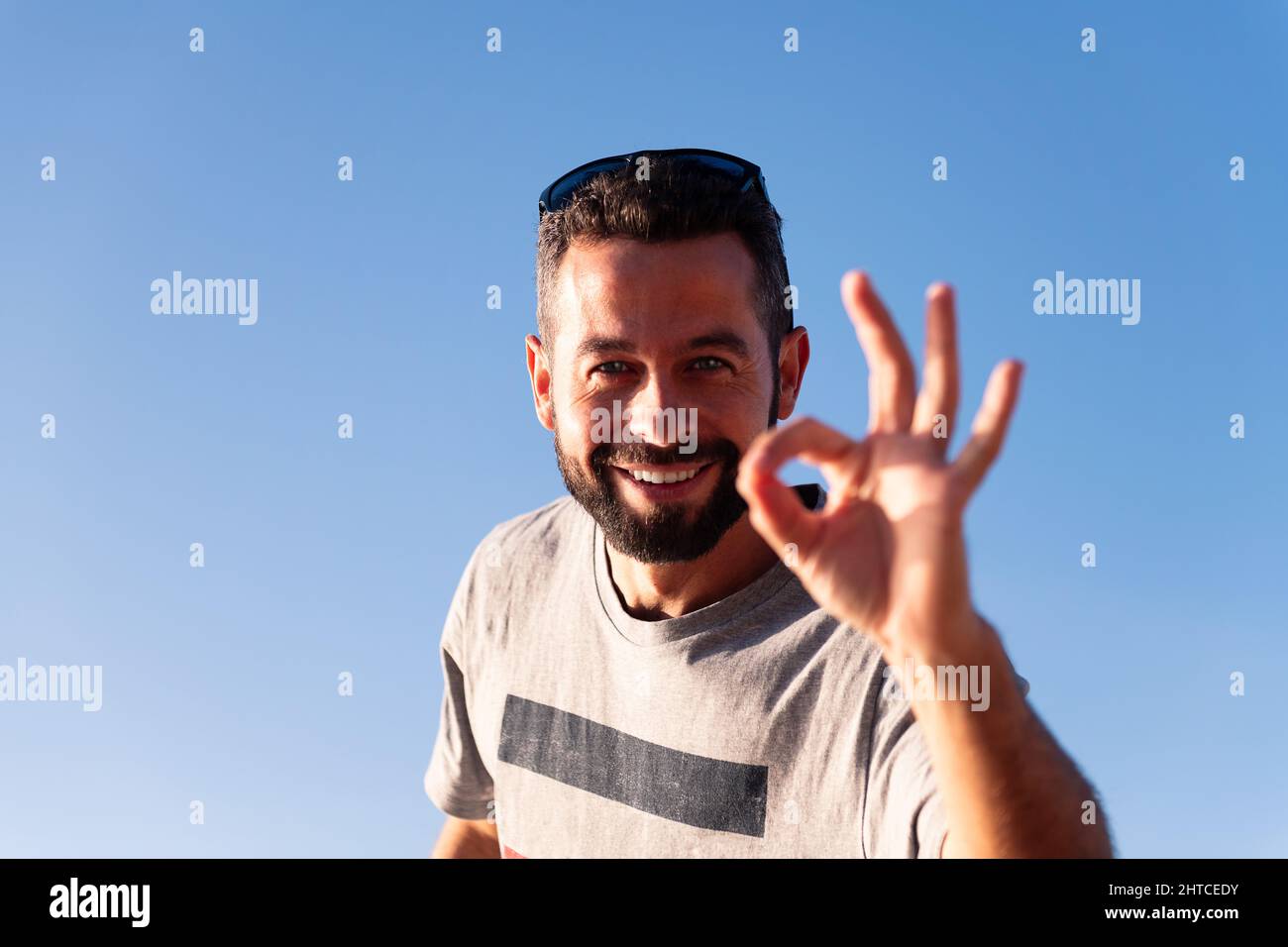 Bärtiger lächelnder Mann, der die Kamera anschaut und das OK-Symbol mit seiner Hand mit dem blauen Himmel im Hintergrund macht, Platz für Text kopieren Stockfoto