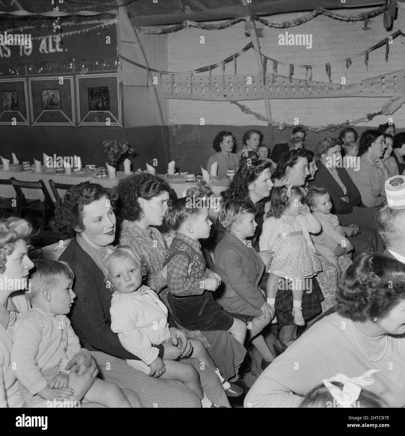 Thurleigh Airfield, Thurleigh, Bedfordfordshire, 19/12/1953. Eltern mit ihren Kindern beobachten die Unterhaltung auf einer Kinderparty. Dieses Foto zeigt eine Kinderparty, die von Laings Wohlfahrtspersonal und Mitgliedern des Komitees für die Kinder von Mitarbeitern organisiert wurde, die am Thurleigh Airfield-Projekt arbeiten. Die Party fand im Camp Theatre statt und beinhaltete Clowns, Spiele, eine Filmshow, Geschenke vom Weihnachtsmann und Tee für sechzig Kinder. Stockfoto