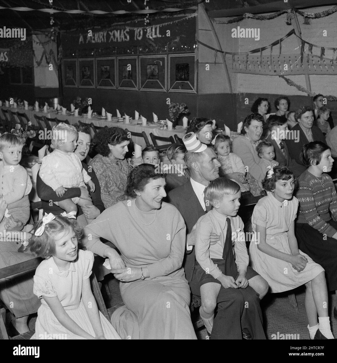 Thurleigh Airfield, Thurleigh, Bedfordfordshire, 19/12/1953. Eltern mit ihren Kindern beobachten die Unterhaltung auf einer Kinderparty. Dieses Foto zeigt eine Kinderparty, die von Laings Wohlfahrtspersonal und Mitgliedern des Komitees für die Kinder von Mitarbeitern organisiert wurde, die am Thurleigh Airfield-Projekt arbeiten. Die Party fand im Camp Theatre statt und beinhaltete Clowns, Spiele, eine Filmshow, Geschenke vom Weihnachtsmann und Tee für sechzig Kinder. Stockfoto