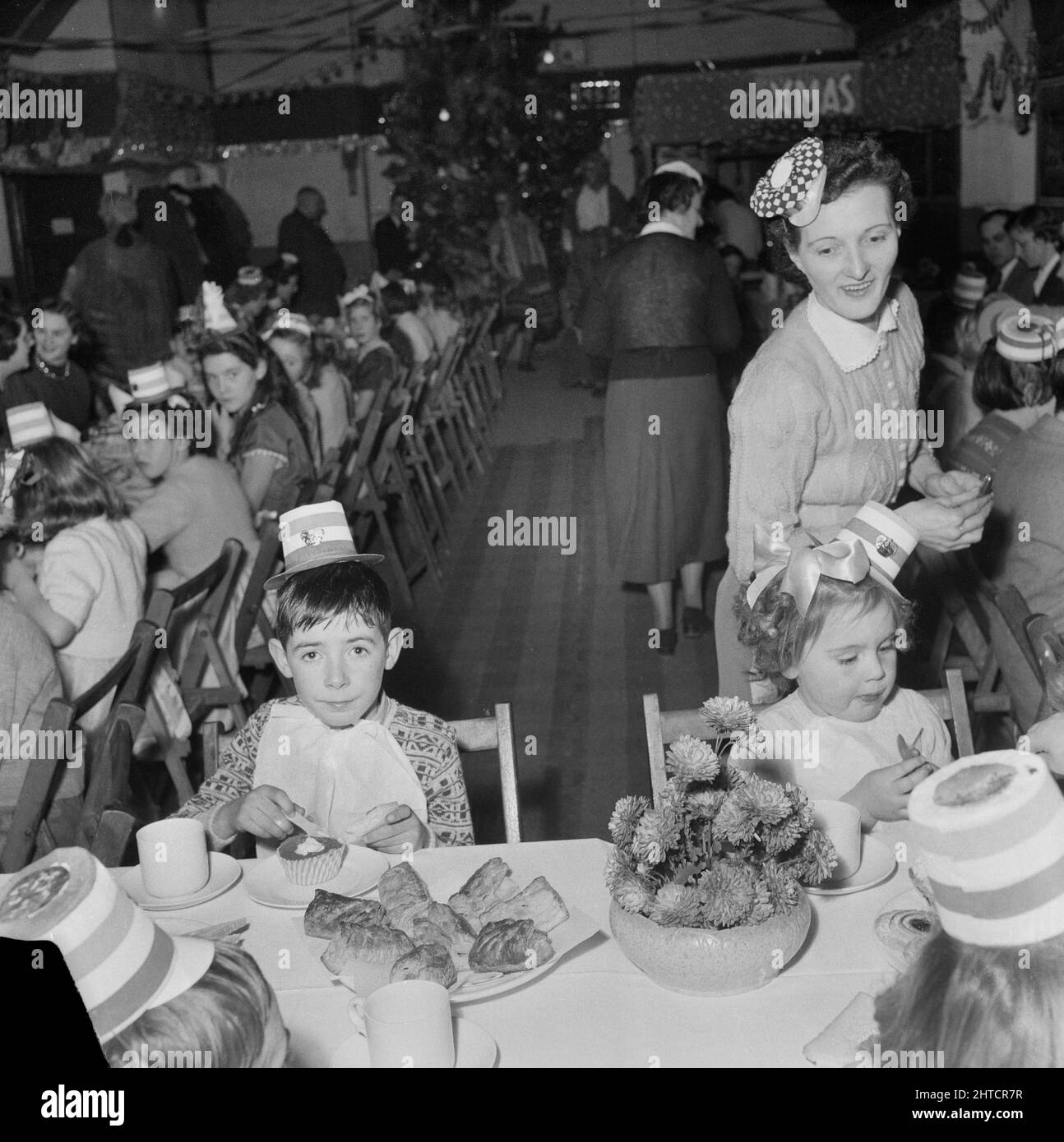 Thurleigh Airfield, Thurleigh, Bedfordfordshire, 19/12/1953. Kinder tragen Partyhüte und haben Tee auf einer Kinderparty. Dieses Foto zeigt eine Kinderparty, die von Laings Wohlfahrtspersonal und Mitgliedern des Komitees für die Kinder von Mitarbeitern organisiert wurde, die am Thurleigh Airfield-Projekt arbeiten. Die Party fand im Camp Theatre statt und beinhaltete Clowns, Spiele, eine Filmshow, Geschenke vom Weihnachtsmann und Tee für sechzig Kinder. Stockfoto