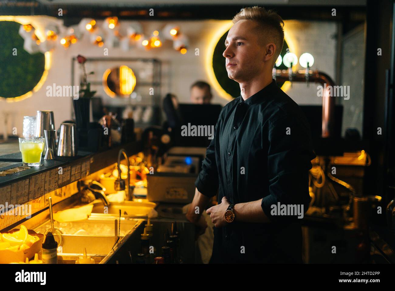 Mittellange Aufnahme eines hübschen jungen Barkeepers, der neben der Theke steht und auf Kundenbestellungen wartet, vor dem Hintergrund eines modernen Nachtclubs. Stockfoto