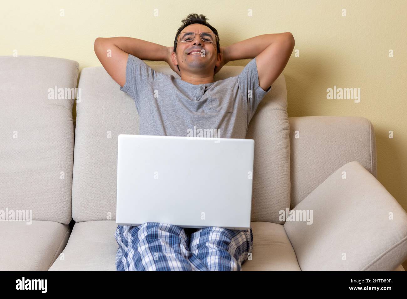 Ein glücklicher, zufriedener junger Mann, der auf einer Couch mit einem Laptop auf den Knien sitzt Stockfoto