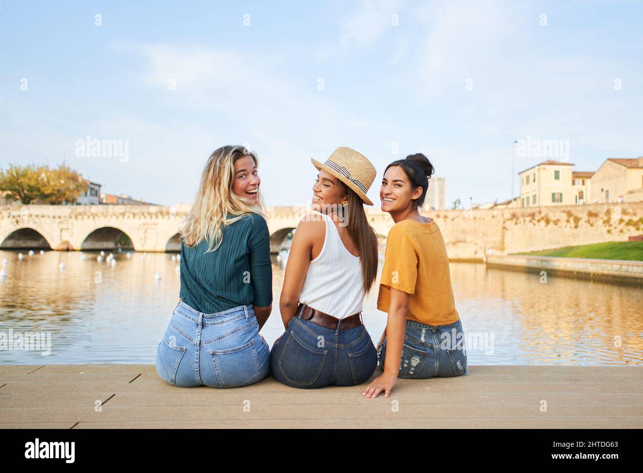 Drei glückliche junge Freundinnen, die Spaß miteinander haben. Eine vielfältige Gruppe von Mädchen, die in der Nähe des Sees der römischen Brückenstadt sitzen. Stockfoto