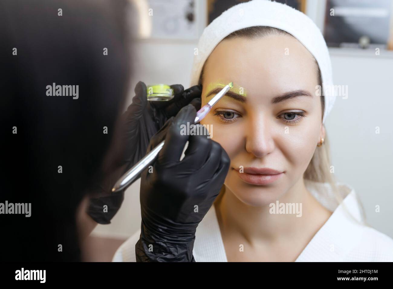 Die Hand eines Make-up Künstlers in schwarzen Handschuhen trägt eine gelbe Augenbrauenpaste auf die Augenbraue auf. Professionelle stilvolle dauerhafte Make-up, Augenbrauen färben Stockfoto