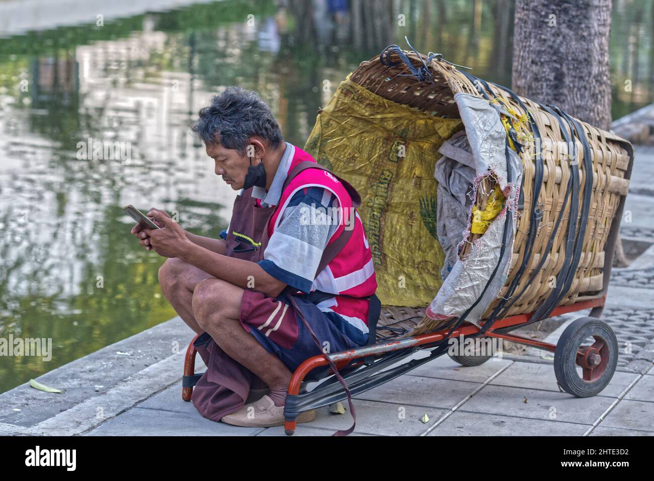 Ein Portier auf einem Markt neben einem langen Kanal in Bangkok, Thailand, sitzt auf seinem umgedrehten Trolley mit seinem Handy Stockfoto