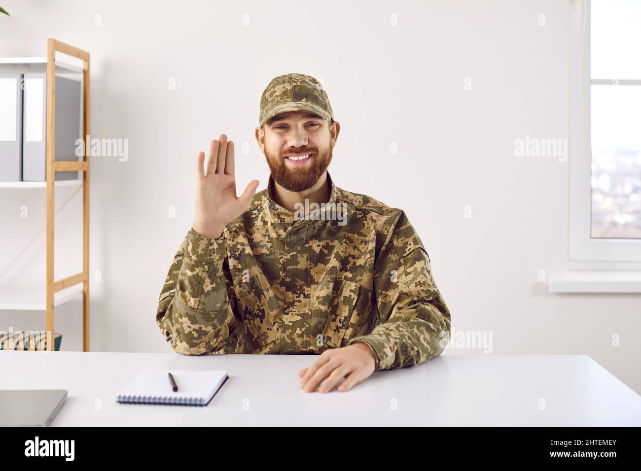 Fröhlicher Soldat, der am Schreibtisch im Büro saß, einen Videoanruf abgab, lächelte und hallo an die Kamera winkte Stockfoto