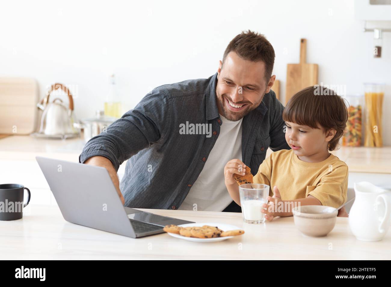 Junger Vater zeigt dem kleinen Sohn auf dem Laptop Lerninhalte, positiver Junge mit gesundem Snack mit Milch in der Küche Stockfoto