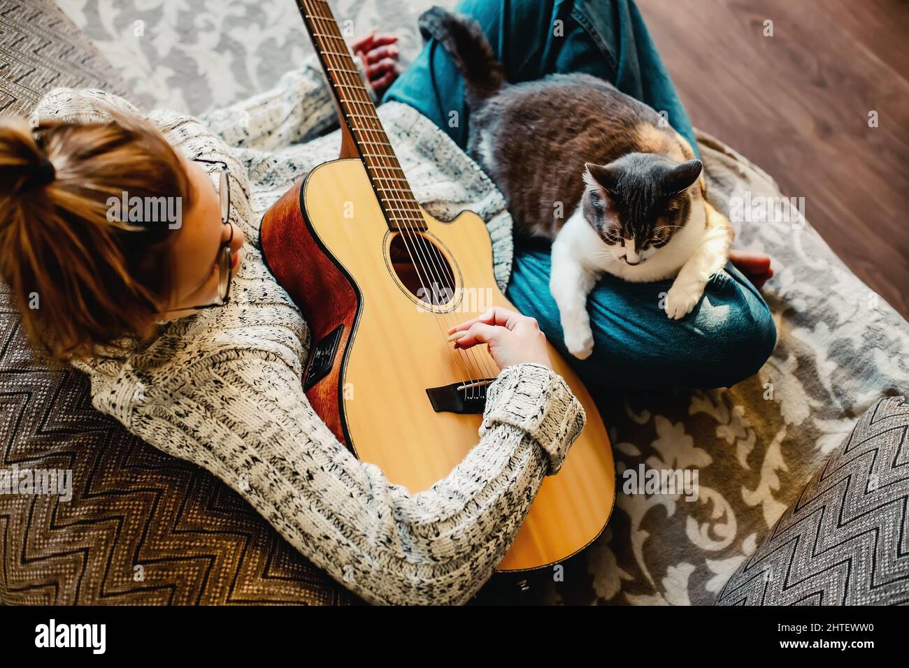 Musikerin sitzt auf der Couch und spielt Gitarre. Katze sitzt auf dem Schoß der Herrin. Spielen Musikinstrument für Haustier. CAT ist Musikliebhaber. Lifestyle. Draufsicht Stockfoto