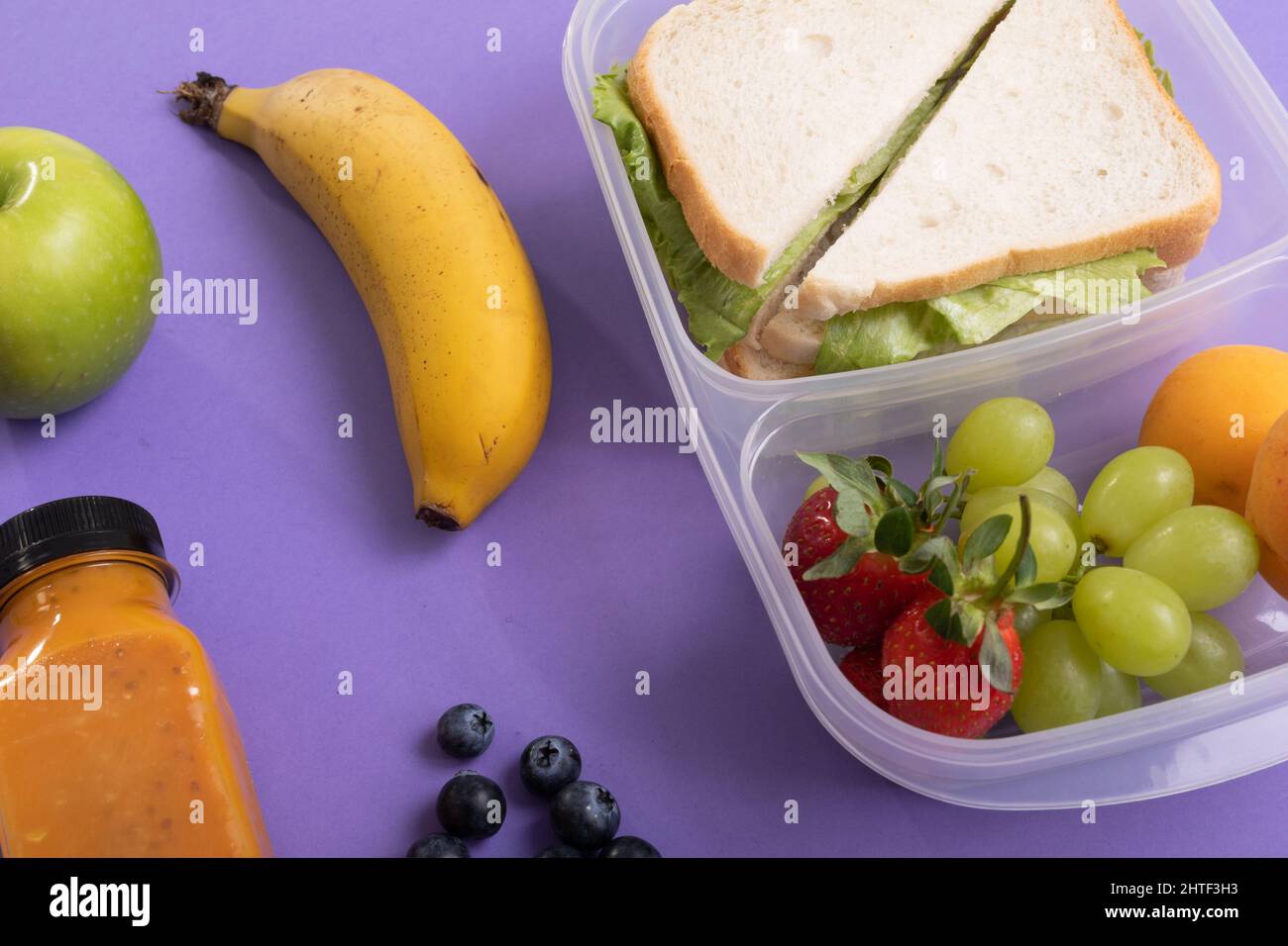 High-Angle-Ansicht von Früchten und Saftflaschen für Sandwiches in einer Tiffin-Box auf violettem Hintergrund Stockfoto