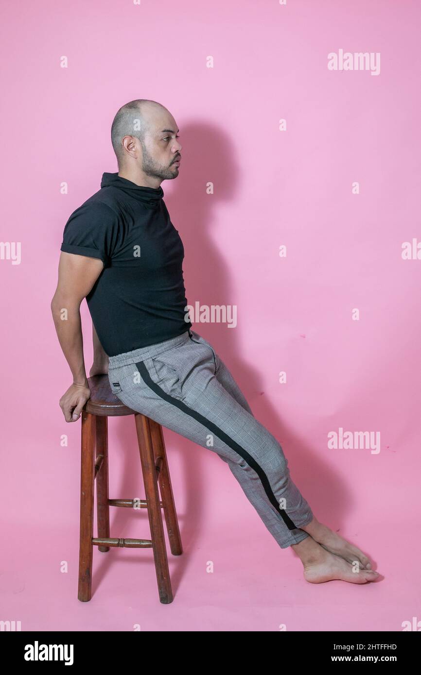 Ganzkörperaufnahme eines jungen, gutaussehenden, kahlen mexikanischen Mannes, der auf einem Stuhl vor einem rosa Hintergrund sitzt Stockfoto